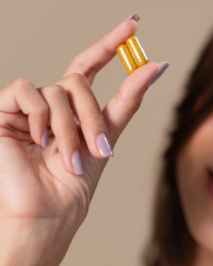 A close-up of a hand holding two small yellow capsules between the fingers against a soft beige background.